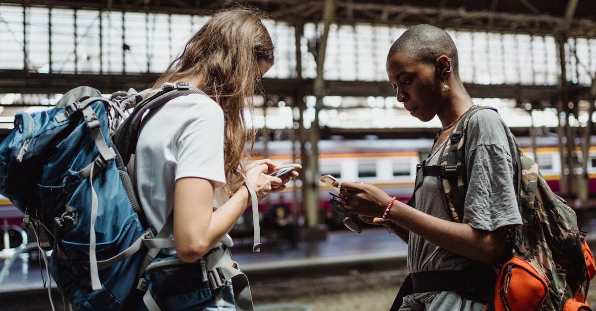 Two backpackers using smartphones at a busy train station preparing for their European journey