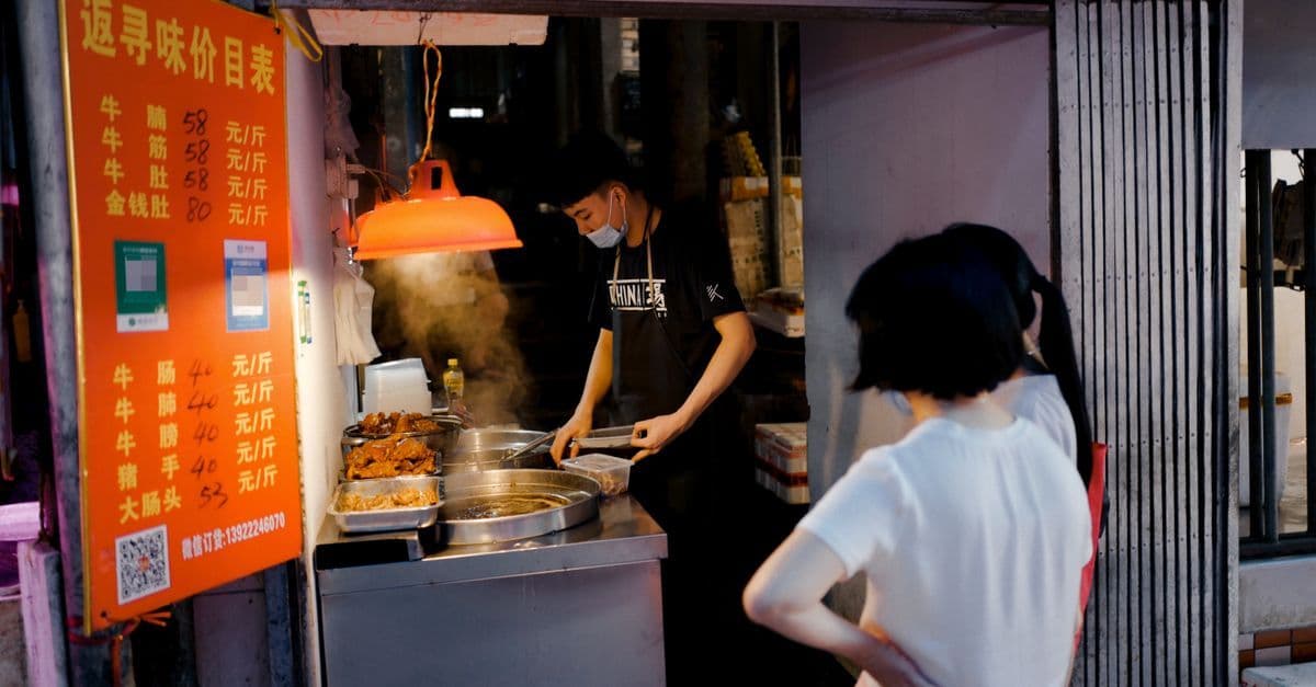 A masked vendor serves food at a Chinese street stall as a customer waits beside the menu.