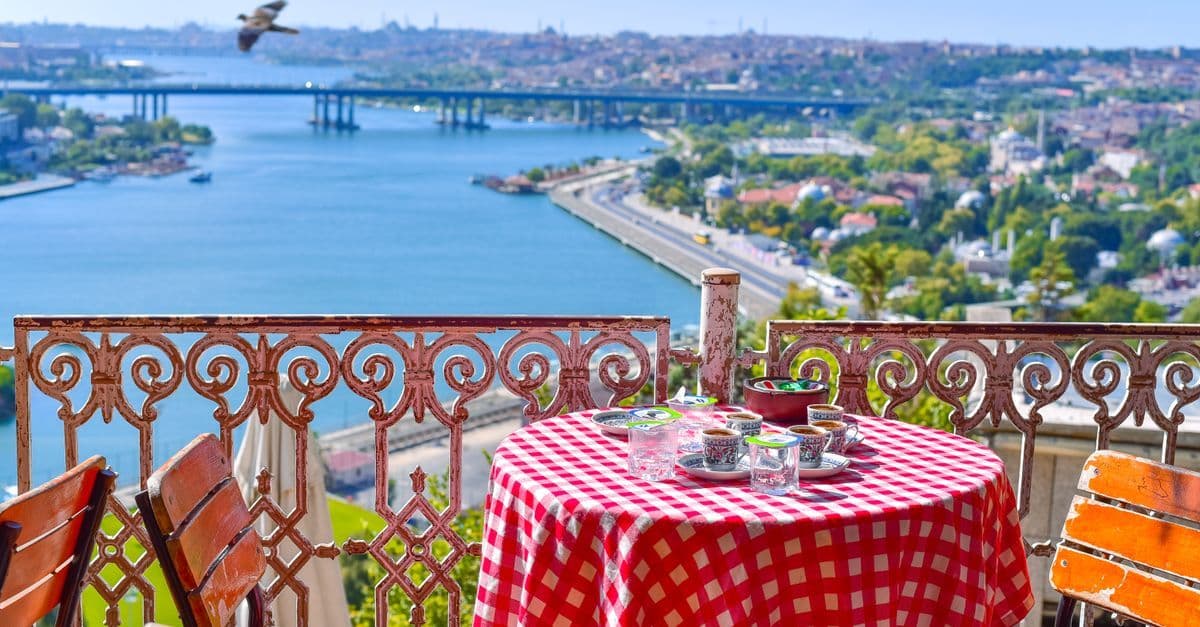 Gedeckter Tisch mit rot-weiß karierter Decke auf einer Terrasse, dahinter Wasser, eine Brücke und die Häuser Istanbuls.
