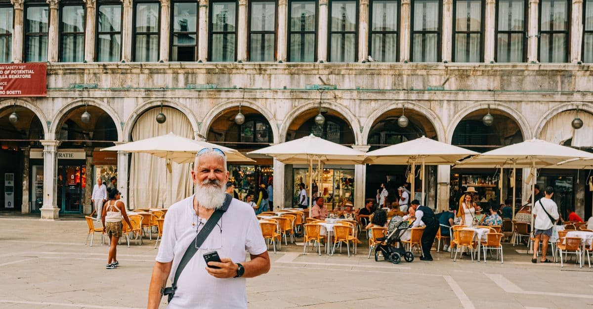 A traveller holds his phone in Piazza San Marco, Venice, in front of a café terrace under white umbrellas.