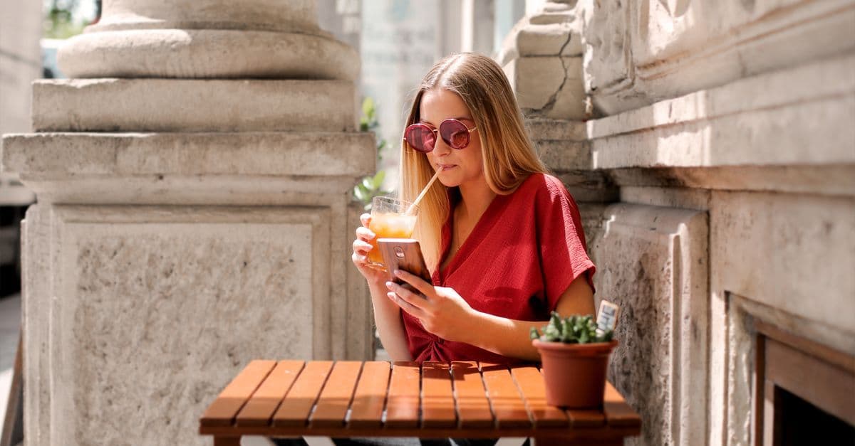 A woman in sunglasses checks her phone while sipping an iced drink at an outdoor cafe beside stone columns.