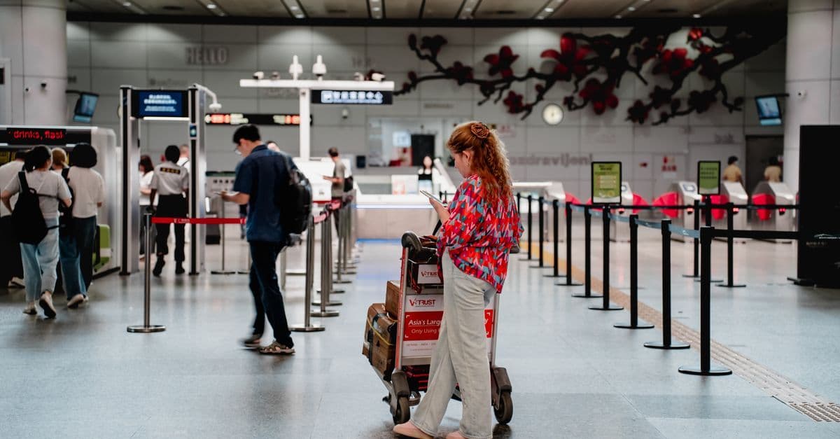 A traveler checks her documents beside a suitcase in an airport hall with counters and queue barriers.