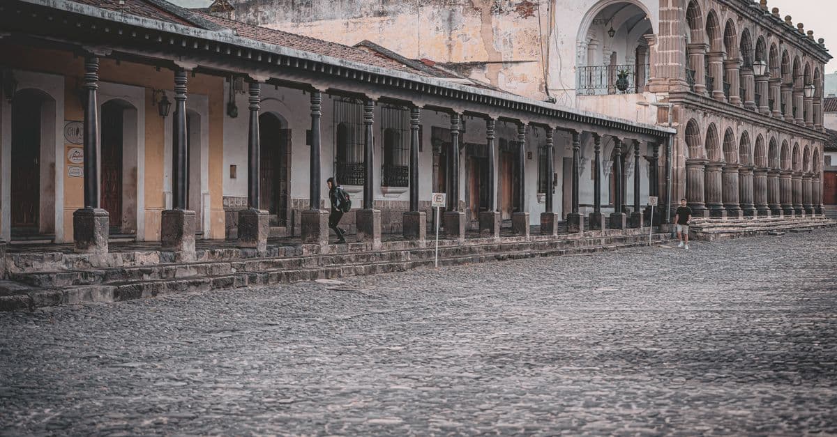 A long colonial building with arched stone columns faces a broad cobblestone street, with a few pedestrians nearby.