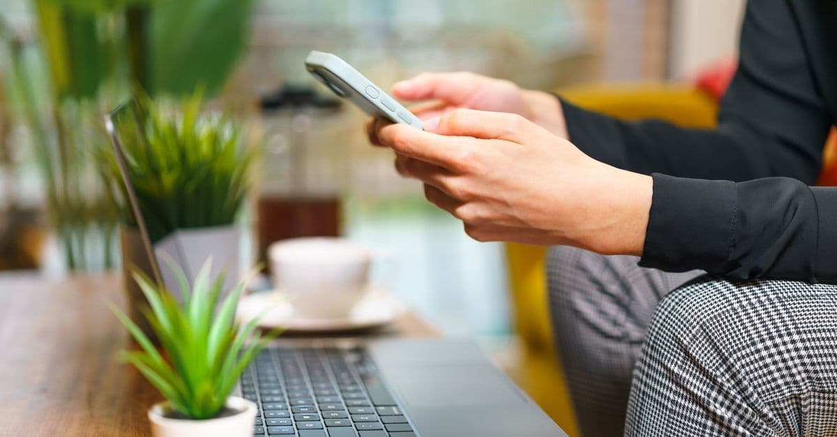 Una persona usa lo smartphone accanto a un portatile, una tazza di caffè e alcune piante.