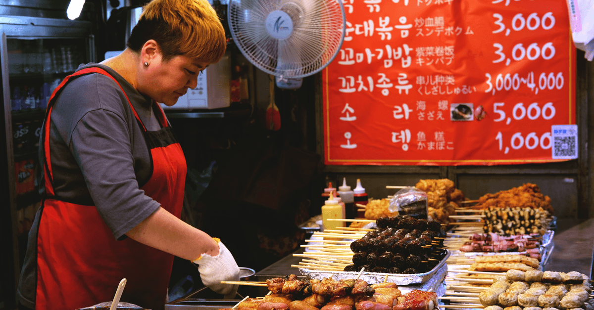 韓国旅行中に屋台で串料理を準備する店主と背景に広がる鮮やかなメニュー看板
