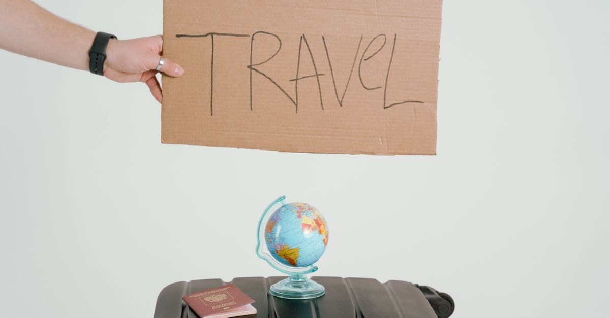Hand hält Pappschild mit Aufschrift 'TRAVEL' über Koffer mit kleinem Globus und Reisepass.