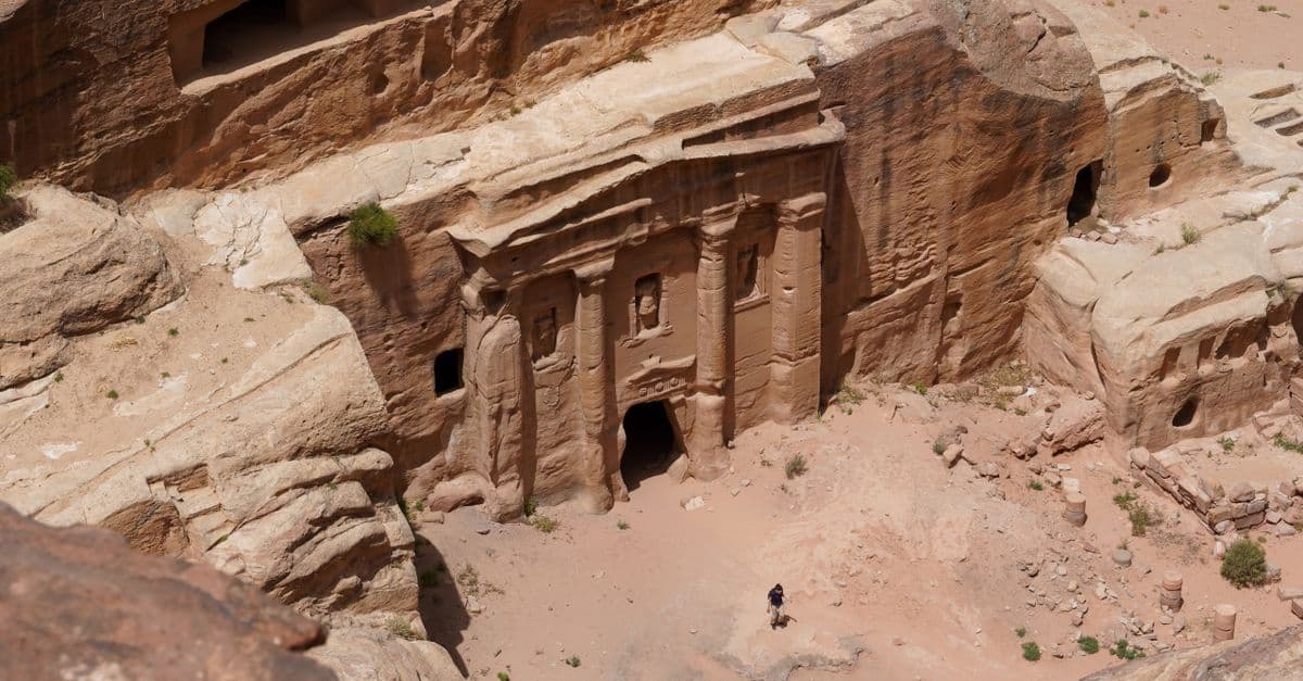 A person stands before an ancient rock-carved tomb in Petra, surrounded by rose-colored sandstone cliffs.