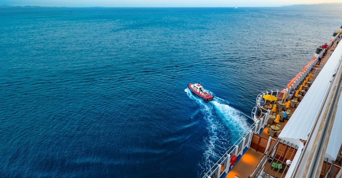 A small boat speeds past a cruise ship on the open sea, seen from one of the ship's upper decks.