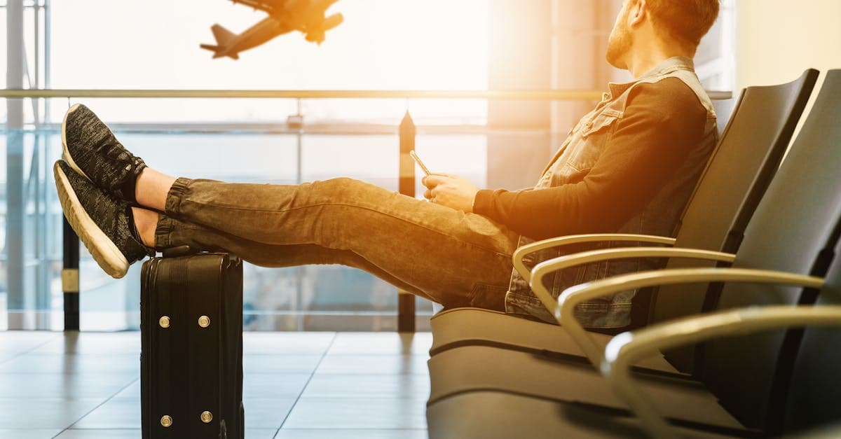 Side view full body of bearded man sitting on chair in waiting hall of airport and looking at flying aircraft behind window