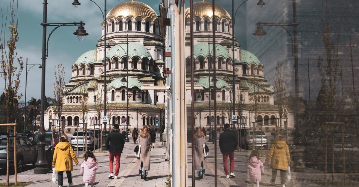 Pedestrians walk past a glass facade reflecting Sofia’s Alexander Nevsky Cathedral.