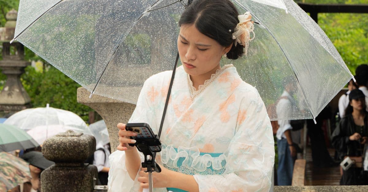 A woman in a kimono checks her phone under a clear umbrella on a rainy day in Kyoto, Japan.