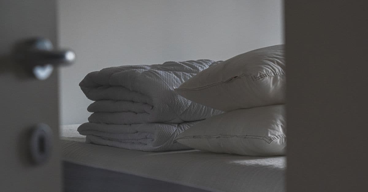 Gestapeld beddengoed met kussens en een dekbed zonder overtrek op een opgemaakt bed gezien door een open deur.