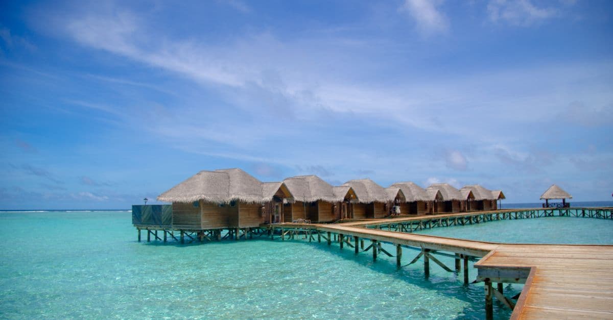 Reihen von Stelzenbungalows stehen über türkisblauem Wasser und sind durch einen Holzsteg mit dem Ufer verbunden.