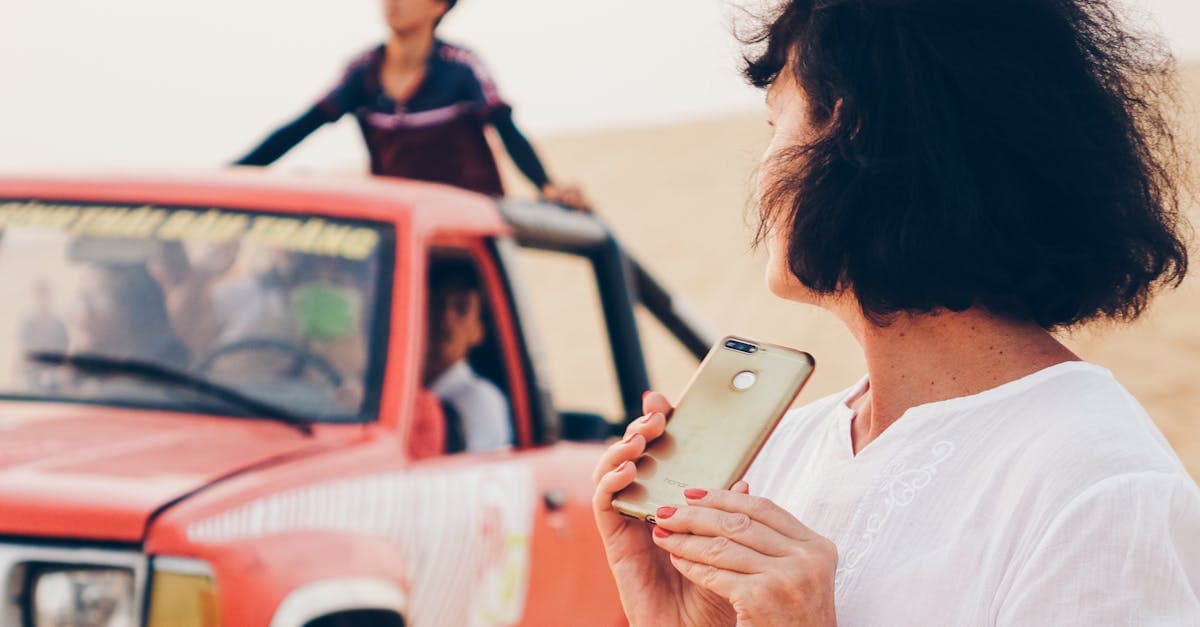 A woman stands in a desert holding a smartphone beside a red off-road vehicle with people.