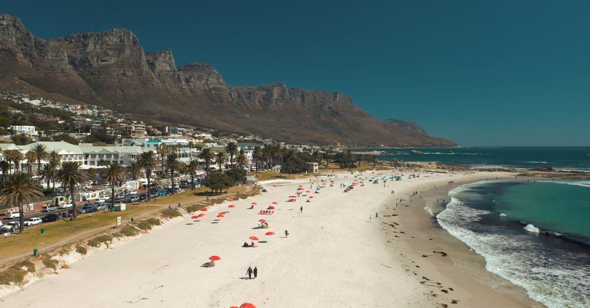A wide sandy beach with scattered umbrellas and swimmers lies beneath a mountain and coastal road in Cape Town.