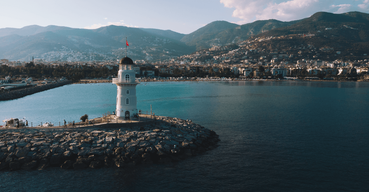 Ein Leuchtturm auf einer Mole an der türkischen Küste, dahinter liegen Stadt und Berge.