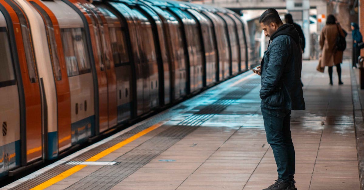 Man texting on his phone while waiting at a railway platform as a train approaches during daylight.