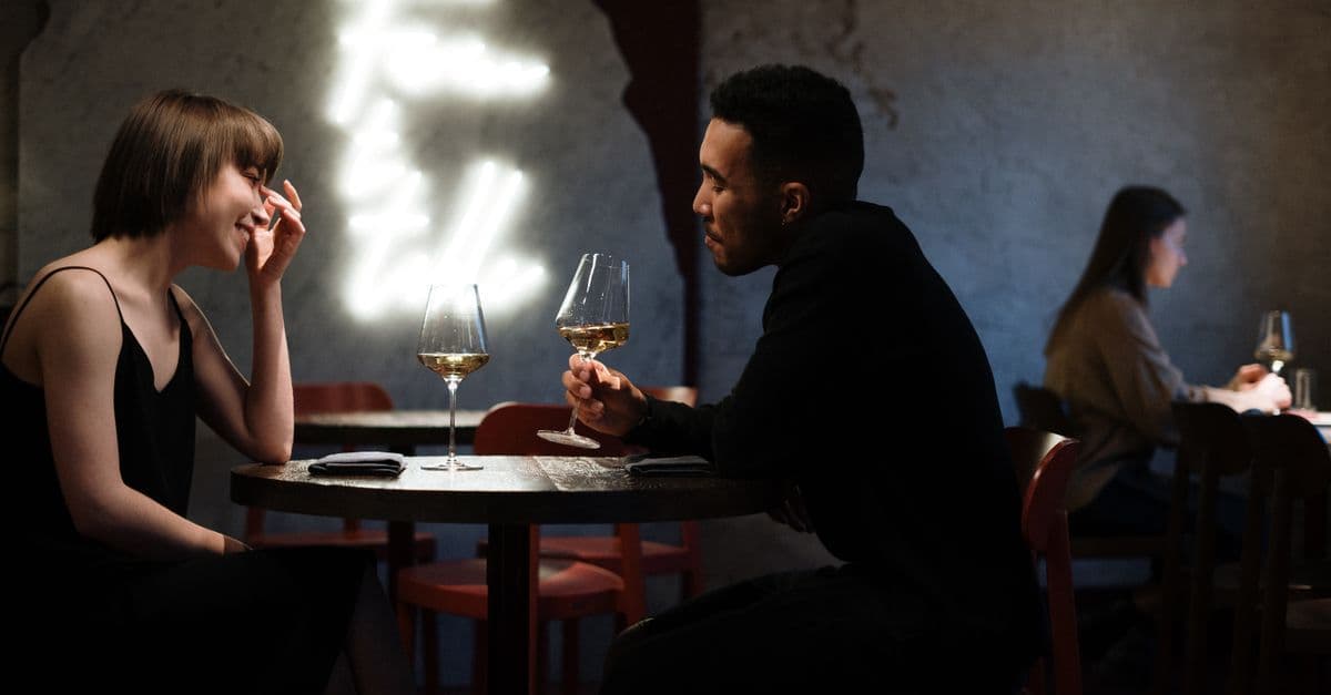 Two people seated at a dim restaurant table smiling and toasting with wine glasses under a soft neon wall light.