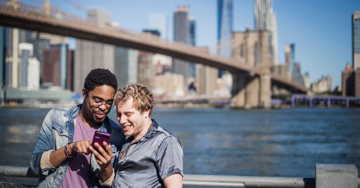 Two smiling friends browsing a smartphone together with the Brooklyn Bridge and NYC skyline behind them.