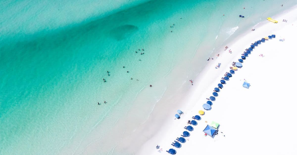 Luchtfoto van wit strand met rij blauwe parasols en turquoise zee waarin mensen zwemmen.
