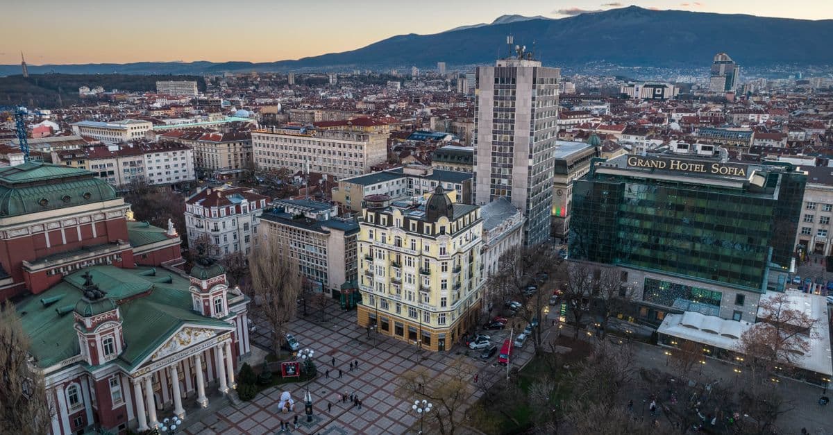 Aerial view of central Sofia, Bulgaria, with historic buildings, a plaza, and mountains beyond.