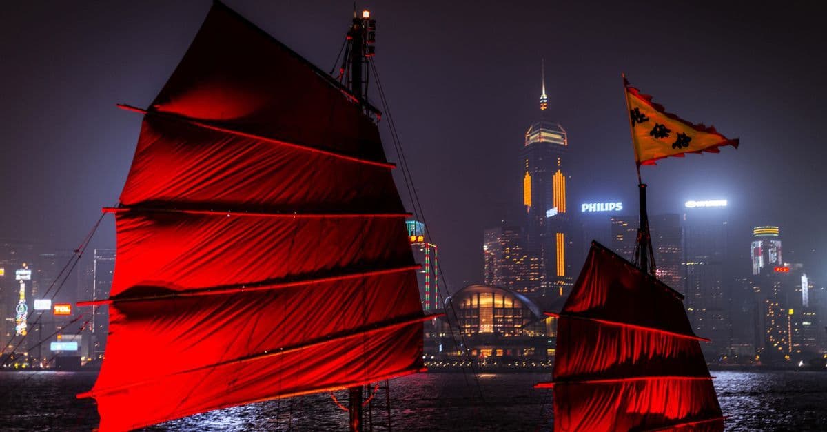 Zwei rot beleuchtete Segelschiffe fahren nachts vor der Skyline von Hongkong.