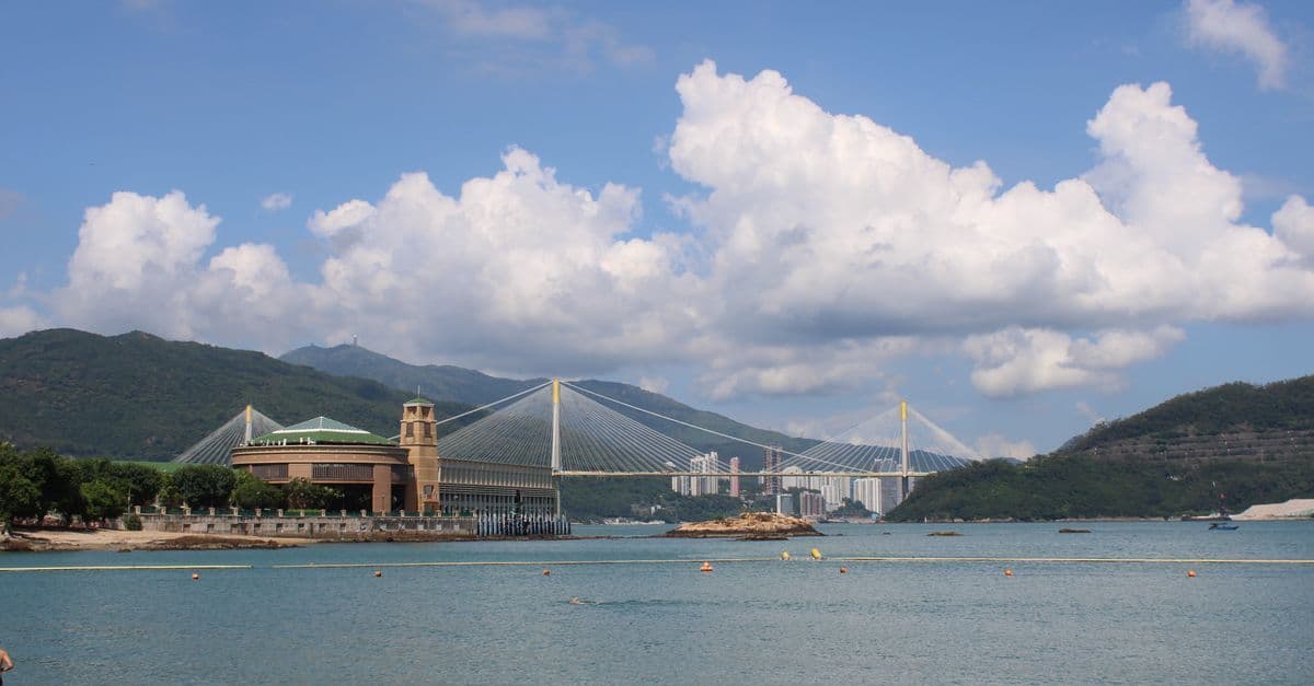 A cable-stayed bridge crosses a bay backed by green hills beneath large white clouds.