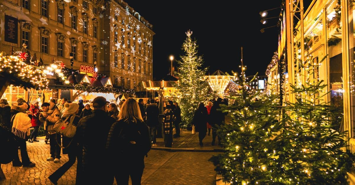 Crowds browse a European Christmas market at night, with glowing stalls and decorated evergreen trees.