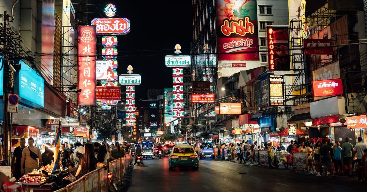 Nächtliche Straße in Bangkok mit leuchtenden Schildern, Essensständen, Passanten und dichtem Verkehr.