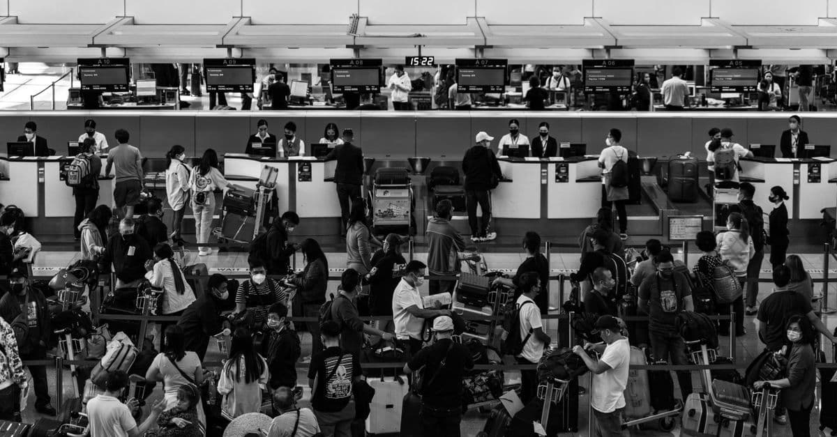 Travelers with luggage queue at airport service counters in a busy terminal.