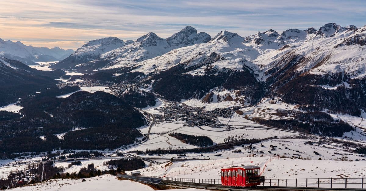 A red mountain railway car climbs above a snowy valley and its villages in the Swiss Alps.