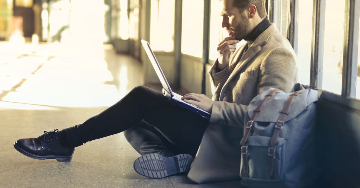A traveller sits on an airport floor by the windows, working on a laptop next to his backpack.