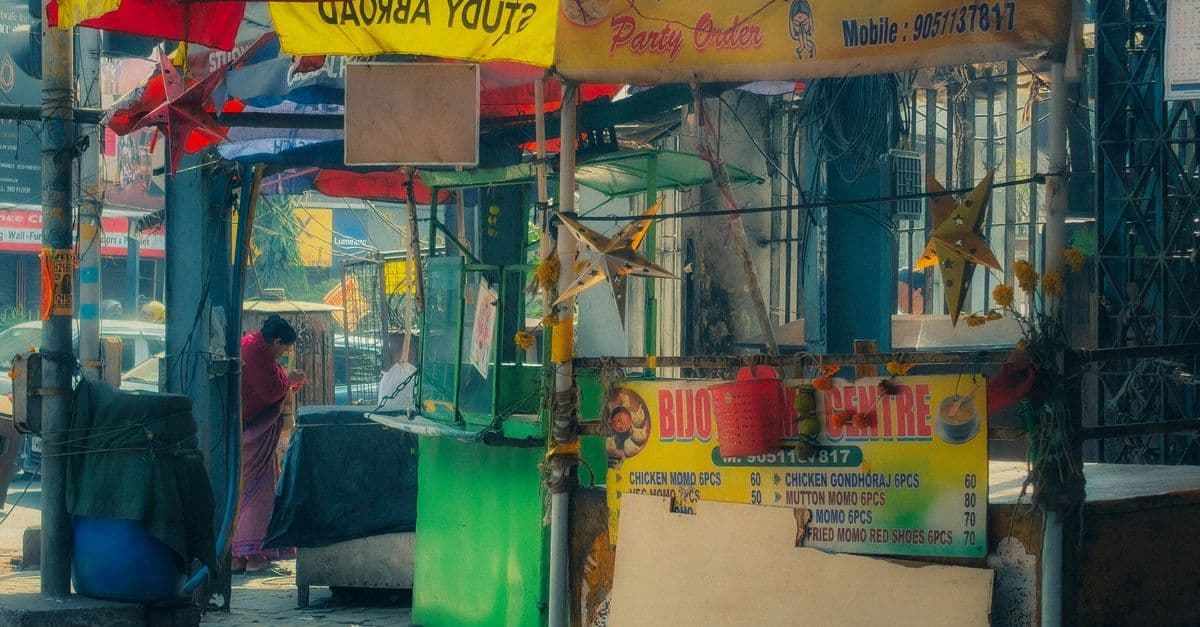 Petit kiosque de rue coloré en Inde, avec un vendeur derrière le comptoir et des enseignes au-dessus.