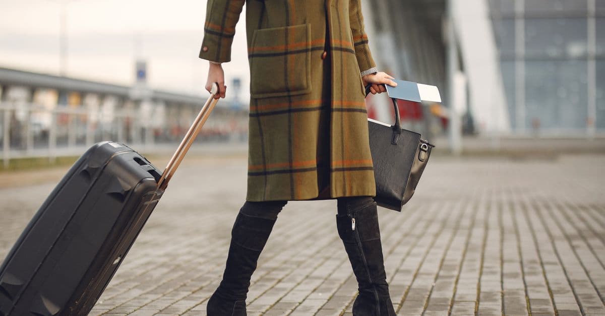 Traveler in a plaid coat pulls a rolling suitcase and carries a handbag outside a modern building.