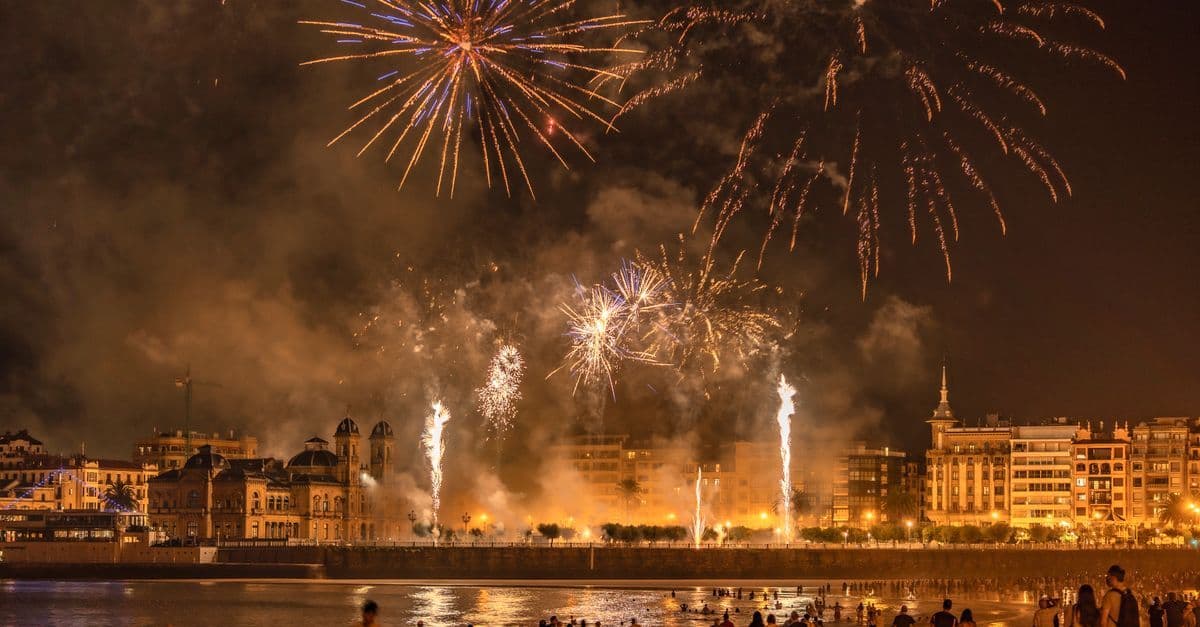 Fireworks light up the night sky above a waterfront city, with crowds watching from the shore.