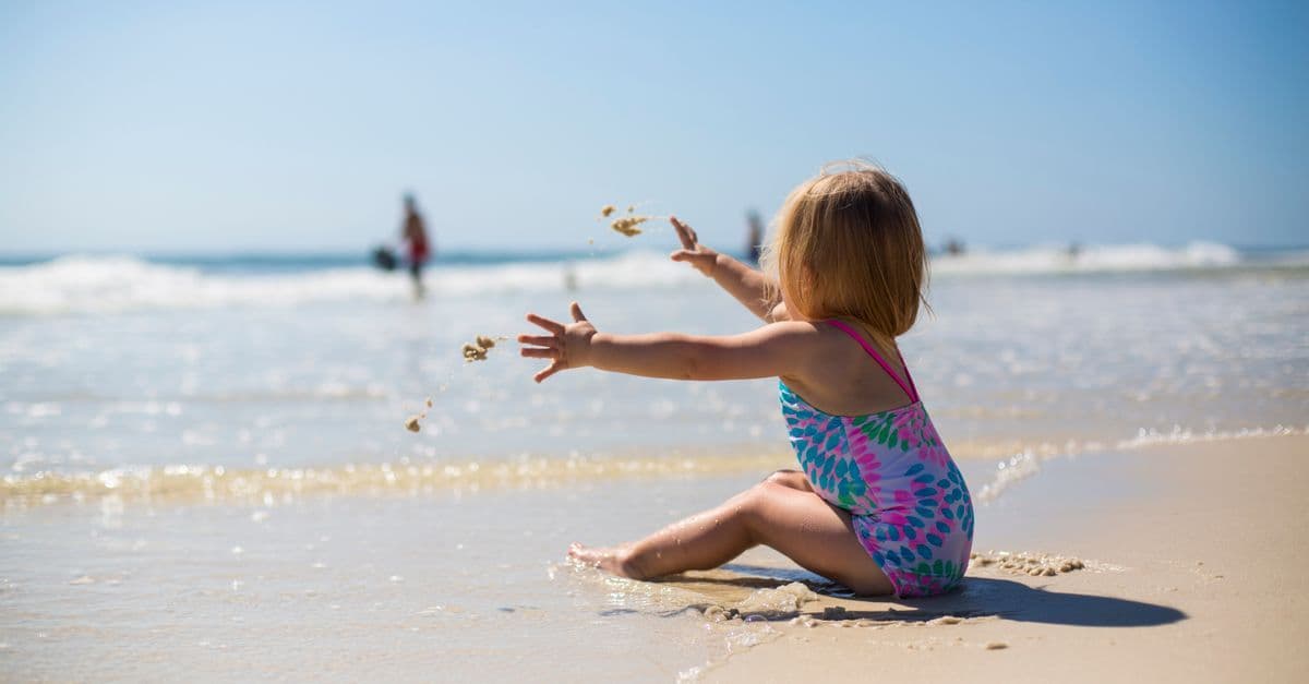 Kleines Kind im bunten Badeanzug sitzt am Strand und wirft Sand ins Meer.