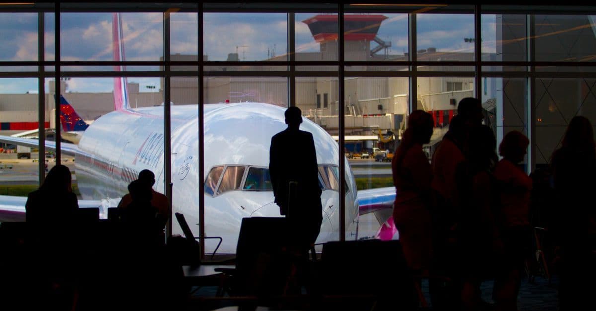 Silhouettes of travelers waiting at an airport terminal with a plane visible through large windows.