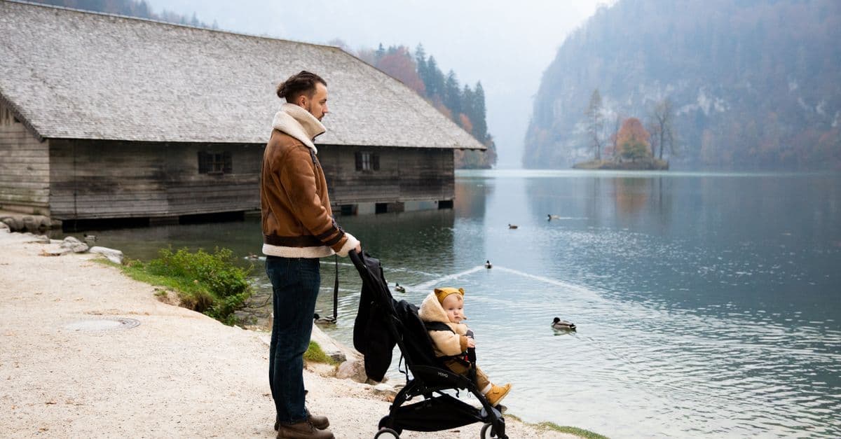 Father pushing a travel stroller beside a peaceful lake with ducks swimming nearby