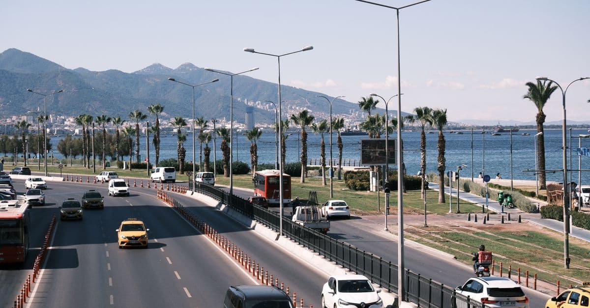 Traffic on a palm-lined seafront road in Izmir, Turkey, with the bay and mountains behind.