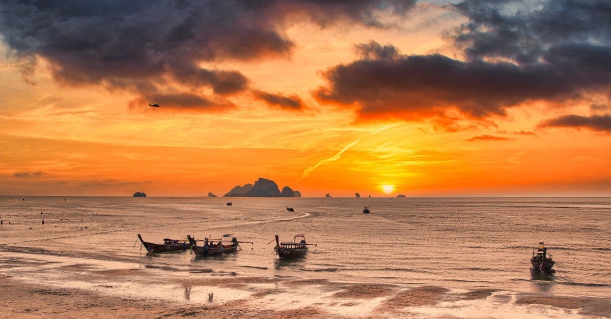 Goldener Sonnenuntergang über dem Strand von Krabi mit traditionellen Longtailbooten und dramatischen Wolken – so schön ist das Thailand-Wetter