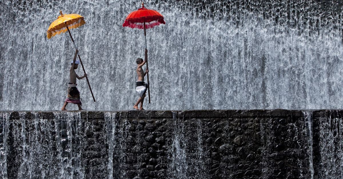 Zwei Männer mit bunten Schirmen in einer Ritualprozession vor einem Wasserfall im Bali Wetter der Regenzeit