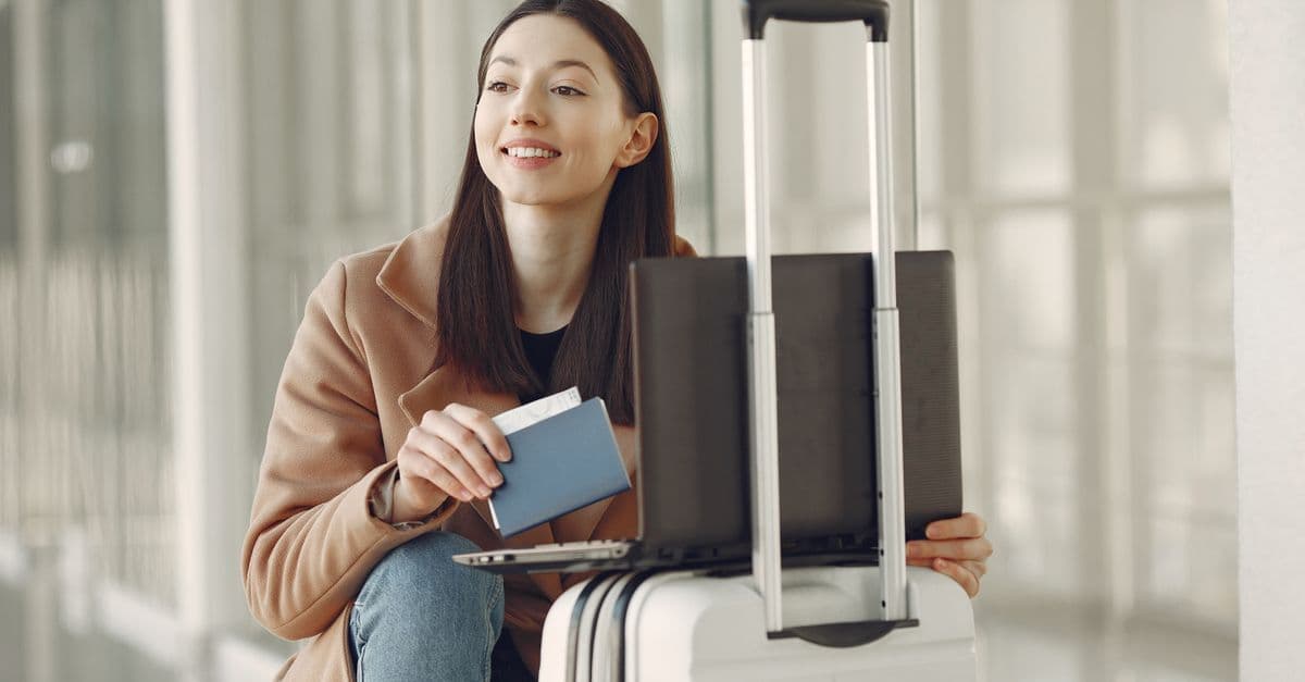 Smiling woman reviewing passport renewal costs on a laptop while seated on luggage in an airport corridor