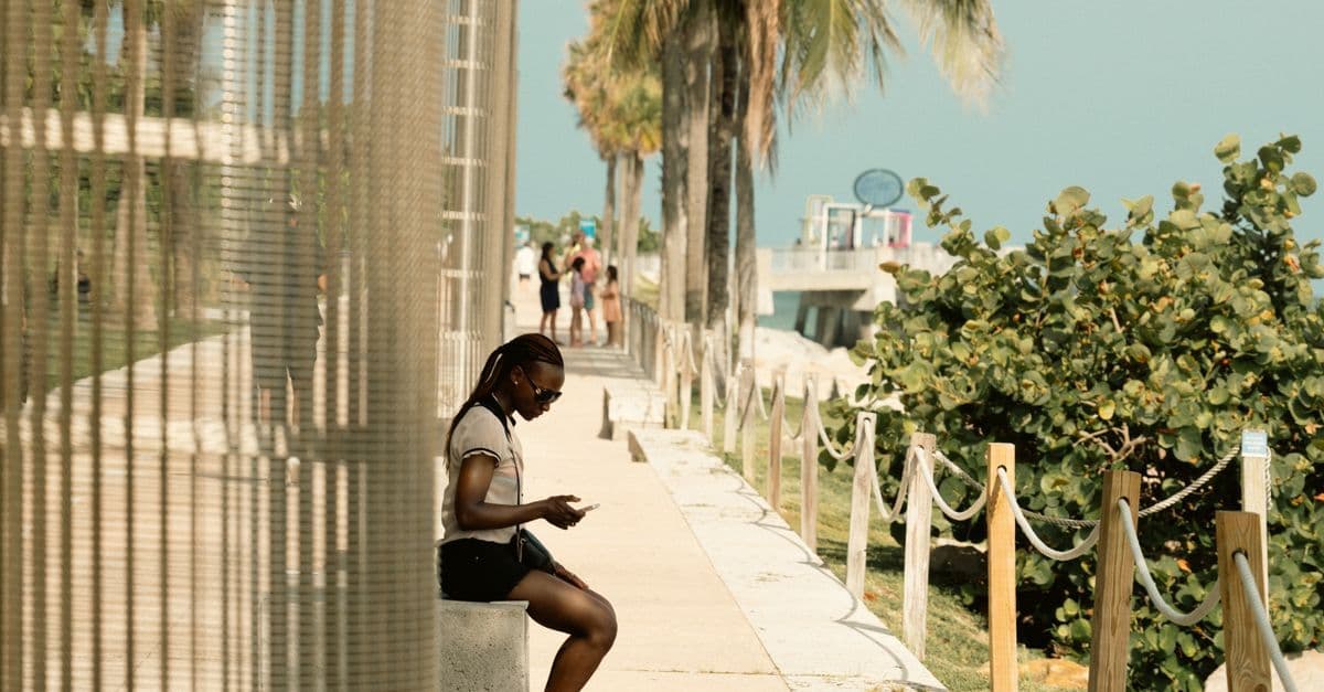 En kvinna sitter på en bänk och använder mobilen vid strandpromenaden i Miami, med palmer längs gången.