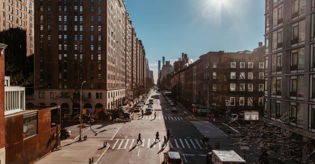 People cross a sunlit avenue in New York City, lined with tall brick apartment buildings and parked cars.