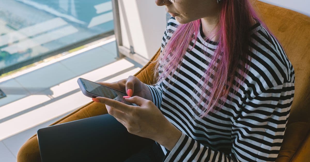 Giovane donna con capelli rosa usa lo smartphone vicino a una finestra, connessa con sky wifi in fibra ottica.