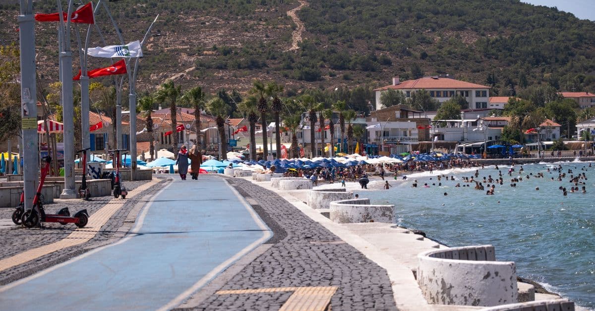 Die Strandpromenade von Foça an der türkischen Ägäis mit Flaggen, Sonnenschirmen und Badegästen im Meer.