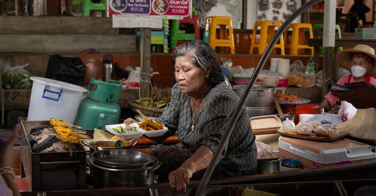 Ältere Thailänderin bereitet frische Streetfood-Gerichte auf einem belebten Bangkoker Markt zu
