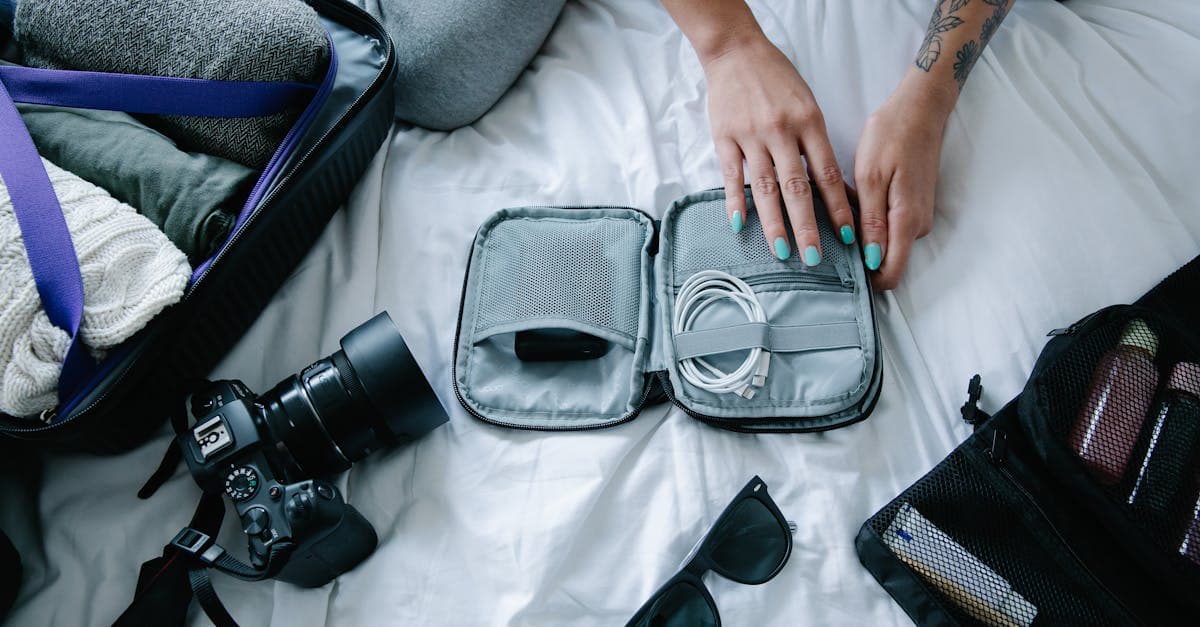Tattooed woman carefully packing electronics, a camera, and sunglasses into a suitcase as carry-on essentials.