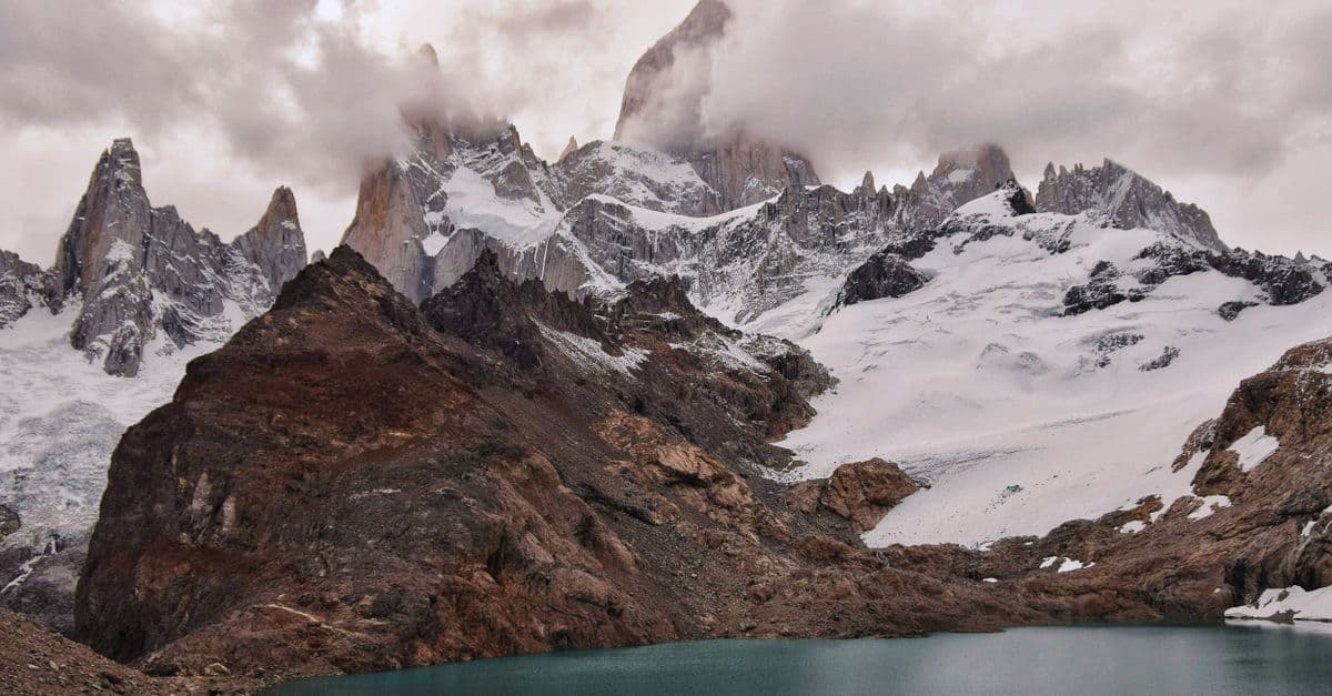 Jagged, snow-covered Patagonia peaks rise above rocky slopes and a turquoise glacial lake.