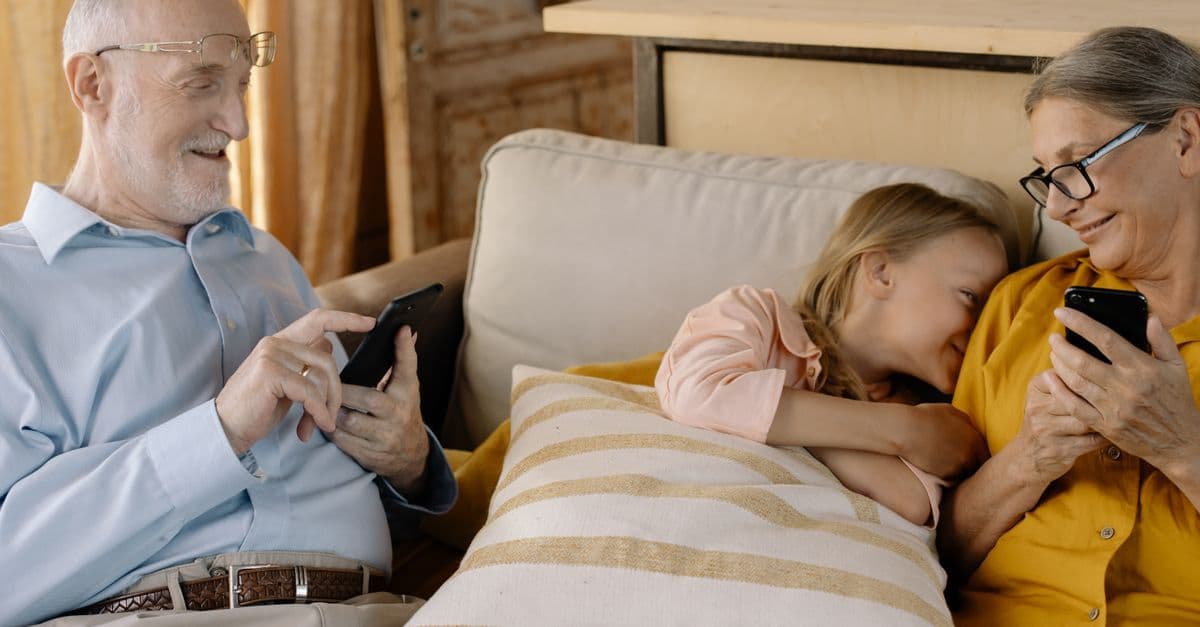 Nonni e nipote sorridenti sul divano mentre usano gli smartphone durante una vacanza in Trentino.